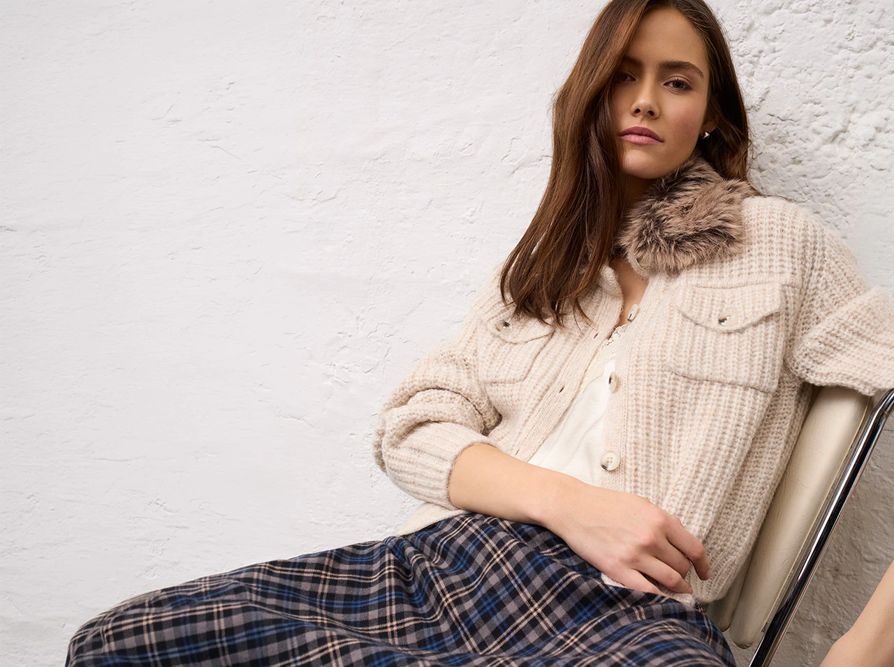 Model trägt eine cremefarbene Strickjacke mit Fellkragen über einem hellen Oberteil und einen blau-braun karierten Rock aus der Oui Heritage Reset Kollektion.