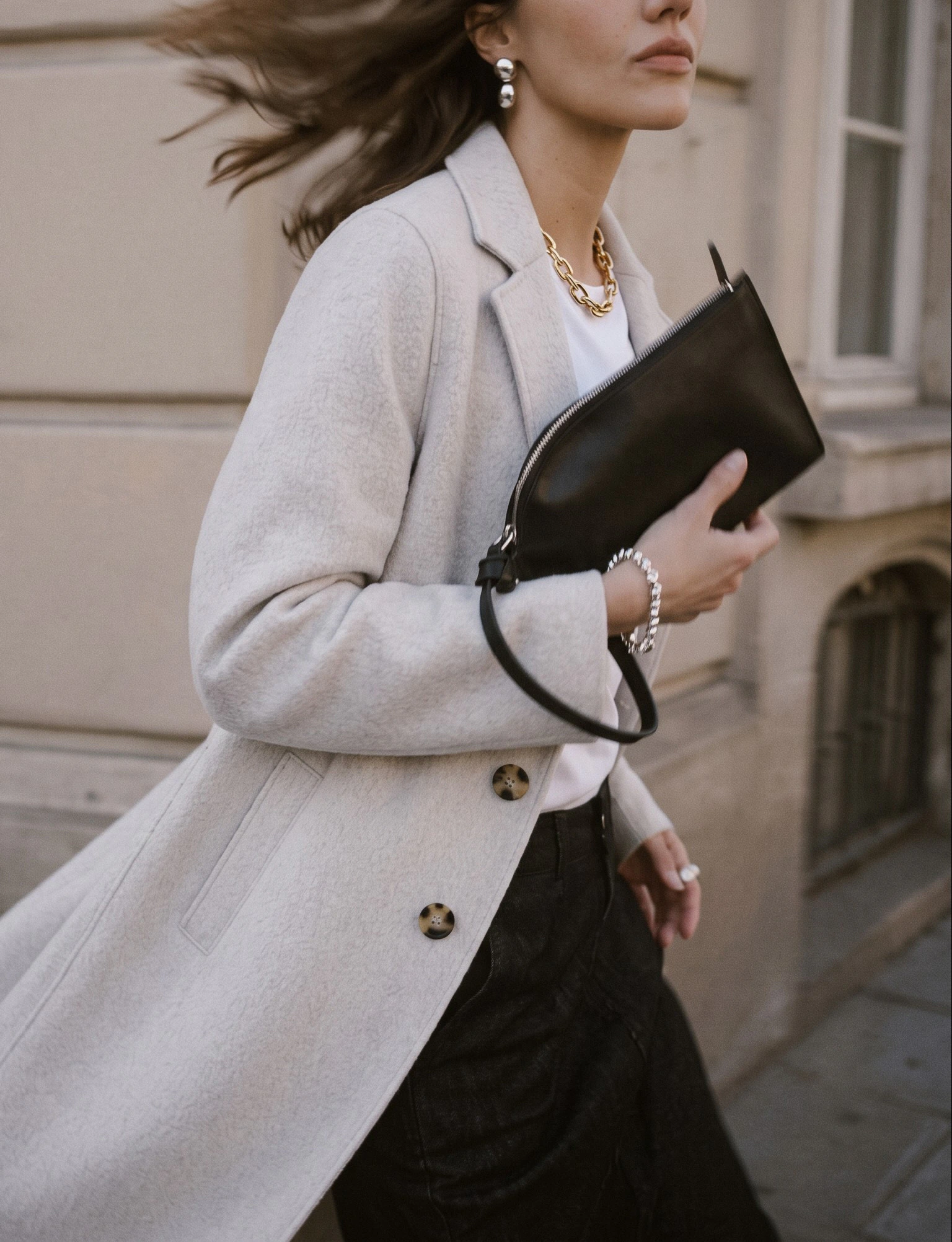 Oui Essentials: Cremefarbener Mantel mit schwarzer Hose, weißem Shirt und schwarzer Clutch im urbanen Look.