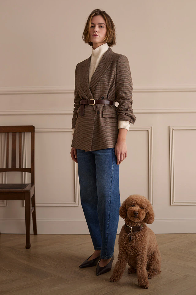 The model is wearing a checked blazer with a belt, a cream-coloured roll-neck jumper and jeans. A brown dog is sitting next to her.