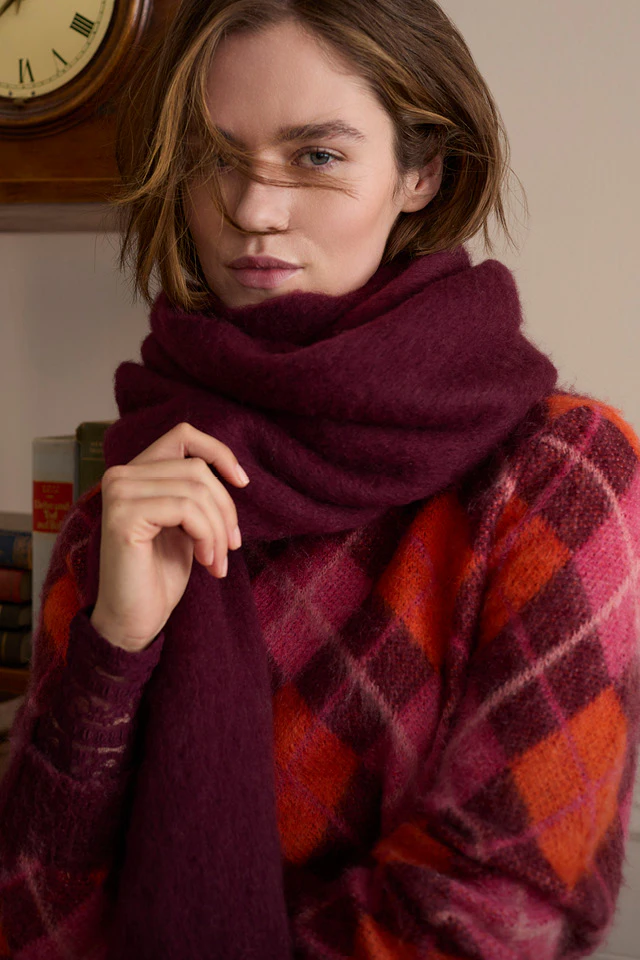 The model is wearing a red and burgundy checked knitted jumper and a voluminous burgundy scarf.