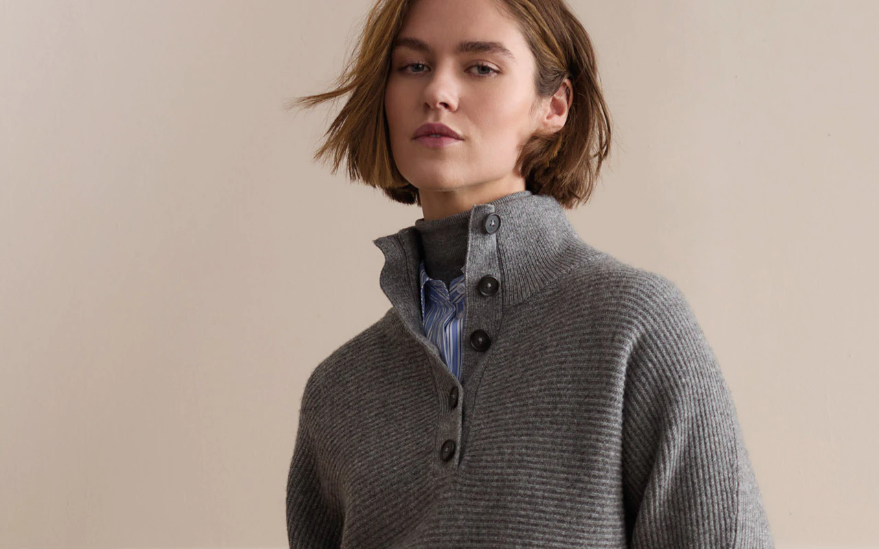 Grauer Strickpullover mit markantem Stehkragen und Knopfleiste, kombiniert mit einer blau-weiß gestreiften Bluse.