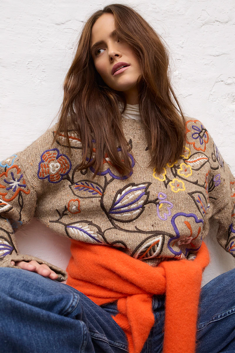 A woman wearing a brown jumper with a floral jacquard pattern, with an orange cardigan tied around her hips, paired with jeans.