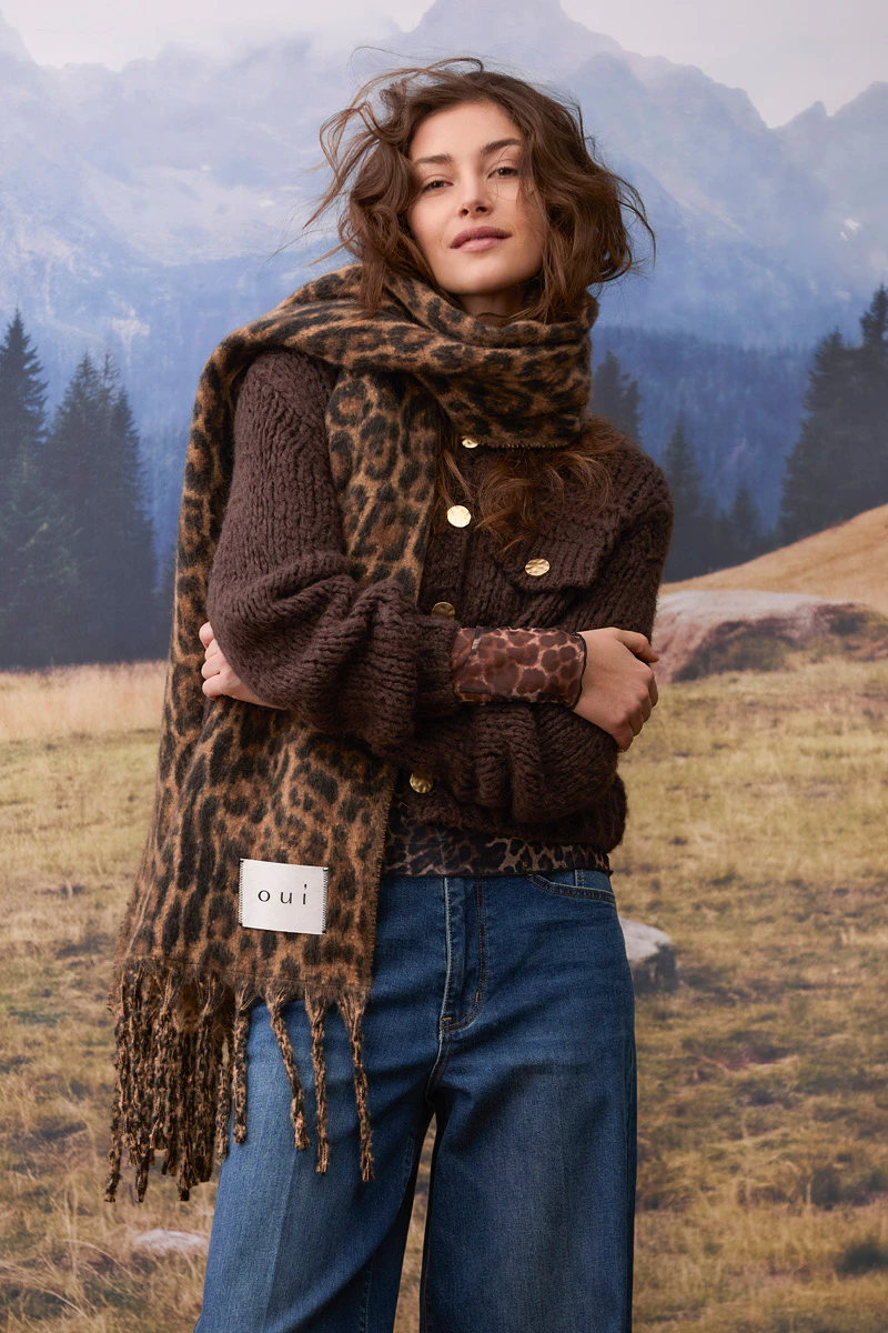 Autumn women’s fashion: a model is wearing a dark brown cardigan with gold-coloured buttons, a leopard-print scarf and a patterned top, paired with dark blue jeans. In the background, an alpine mountain landscape with woods and meadows can be seen.