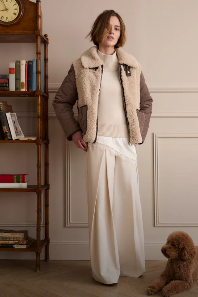 Model trägt eine braune Jacke mit Teddyfutter über einem cremefarbenen Strickpullover und einer weiten cremefarbenen Hose. Neben ihr liegt ein brauner Hund.