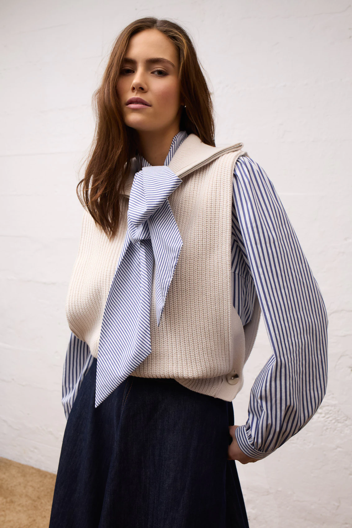 A woman wearing a blue and white striped blouse with a bow, a cream-coloured knitted slipover and a dark blue skirt.