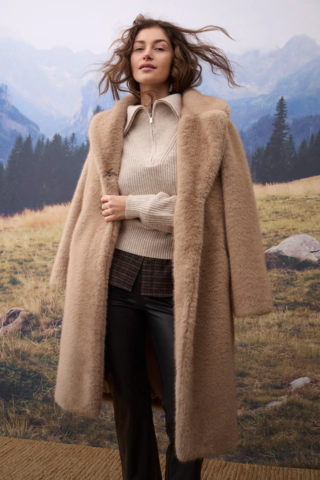 The model is wearing a long beige coat over a light-coloured knitted jumper and a brown checked top, paired with black leather trousers