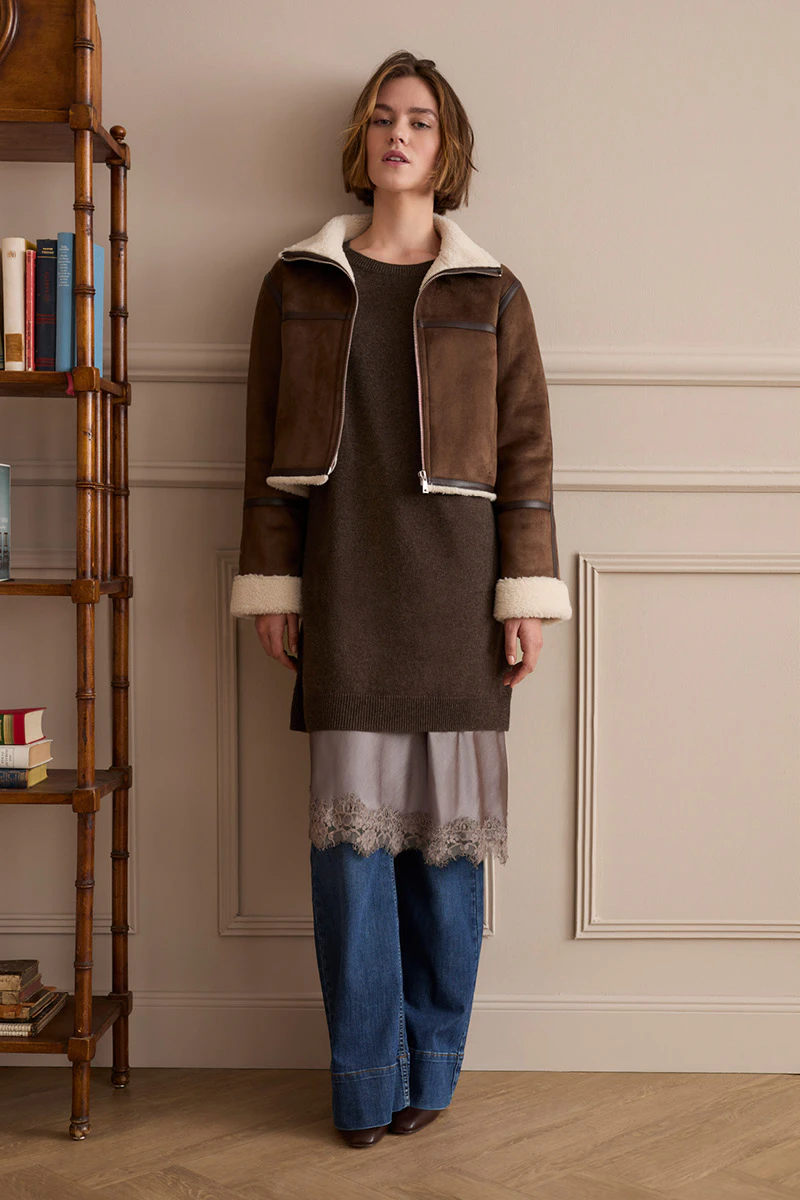 The model is wearing a brown jacket with a teddy bear-print lining over a brown knitted dress and a lace blouse, paired with jeans.