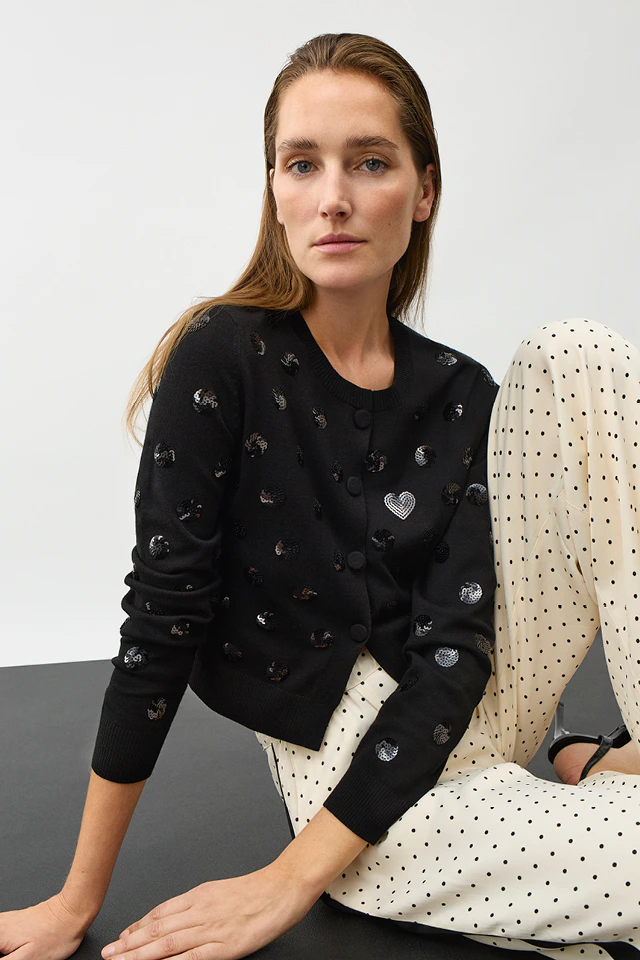 A woman wearing a black cardigan with sequinned dots and white trousers with black dots and black braid down the sides.