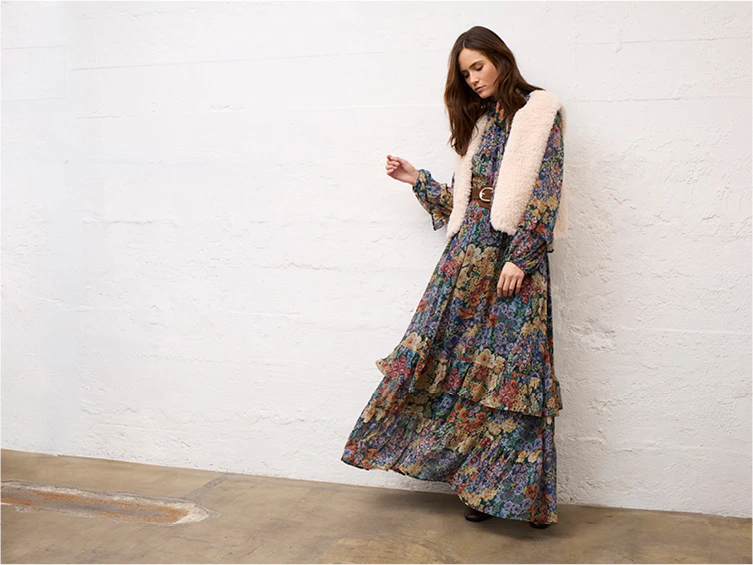 Eine Frau, die ein Midi-Kleid mit Blumenmuster und eine beige Weste trägt.