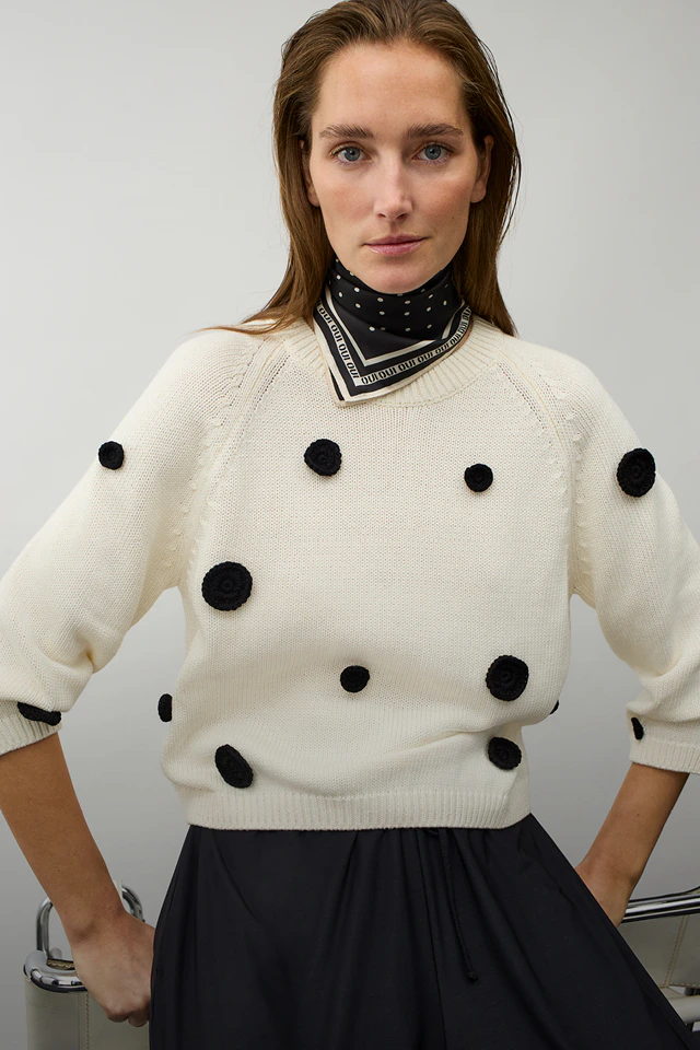 Frau mit weißem Pullover mit schwarzen Punkten, schwarz-weissem Halstuch und schwarzem Rock.