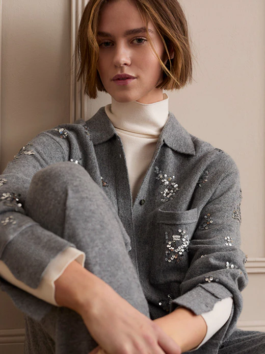 The model is wearing a grey sequinned cardigan over a cream-coloured roll-neck jumper and grey trousers.