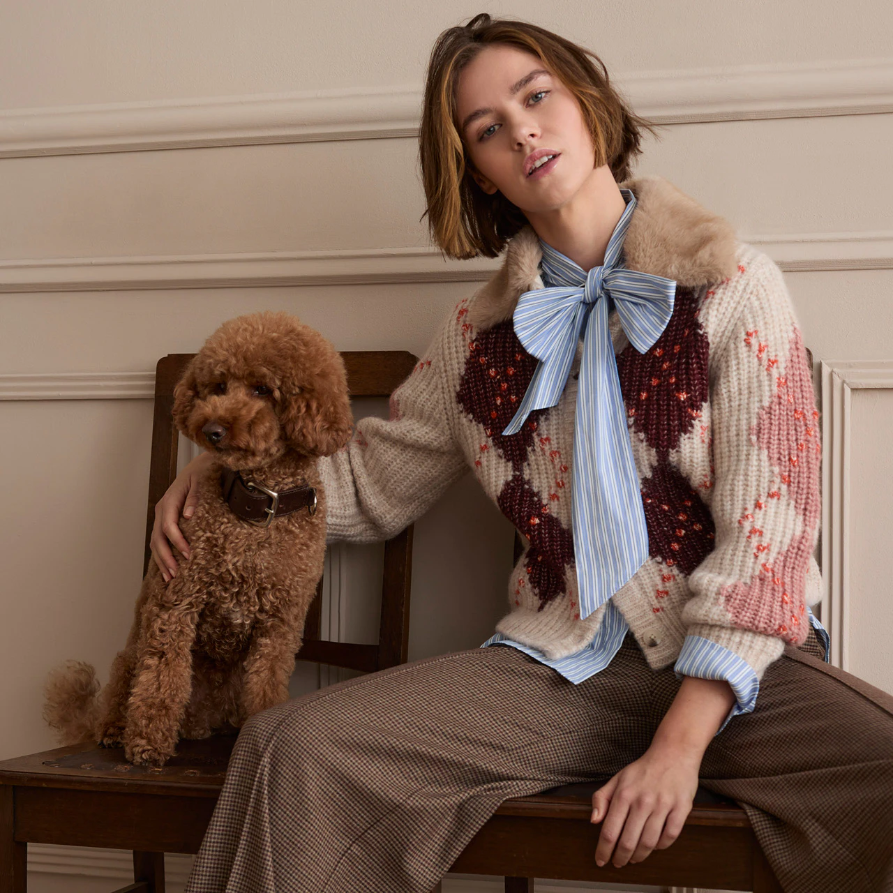 The model is wearing a patterned knitted jumper over a striped blouse with a bow and a checked skirt. A brown dog is sitting next to her.
