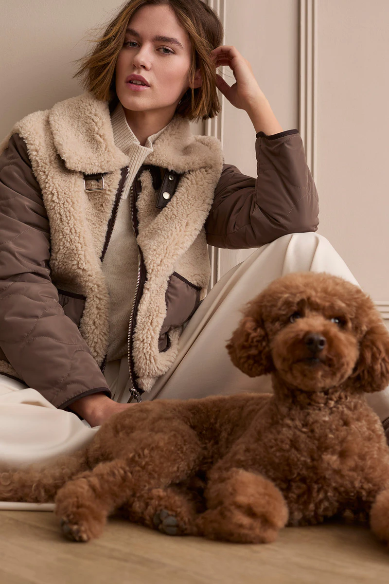 Model trägt eine braune Jacke mit Teddyfutter, cremefarbenen Strickpullover und helle Hose. Im Vordergrund liegt ein brauner Hund.
