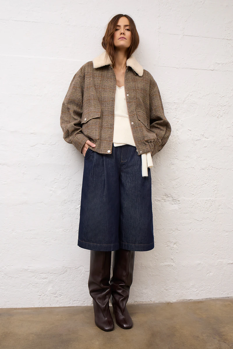 Frau mit brauner Jacke mit weißem plüschkragen, weißem Pullover und dunkelblauer Jeans im kürzeren Schnitt.