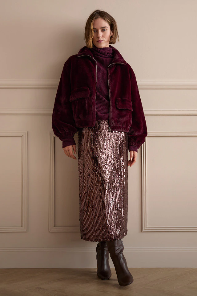 The model is wearing a burgundy jacket and a matching knitted jumper, paired with a shimmering sequinned skirt and boots.