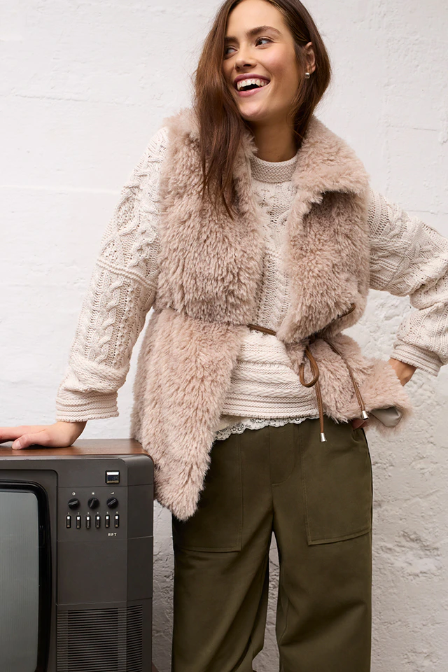 A woman in a cream-coloured knitted jumper, a beige-brown fur waistcoat and loose-fitting olive-green trousers, standing next to a retro television, smiling.