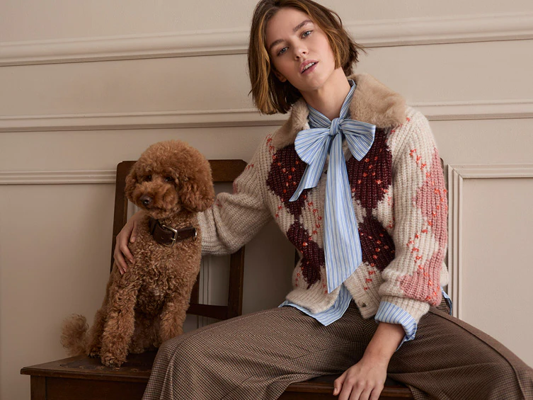 Model trägt einen gemusterten Strickpullover über einer gestreiften Bluse mit Schleife und einen karierten Rock. Neben ihr sitzt ein brauner Hund.