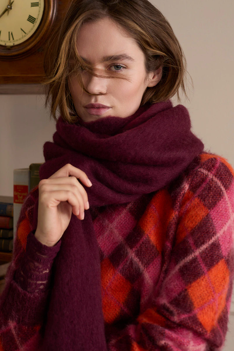 Model trägt einen rot-burgunderfarben karierten Strickpullover und einen voluminösen burgunderfarbenen Schal.
