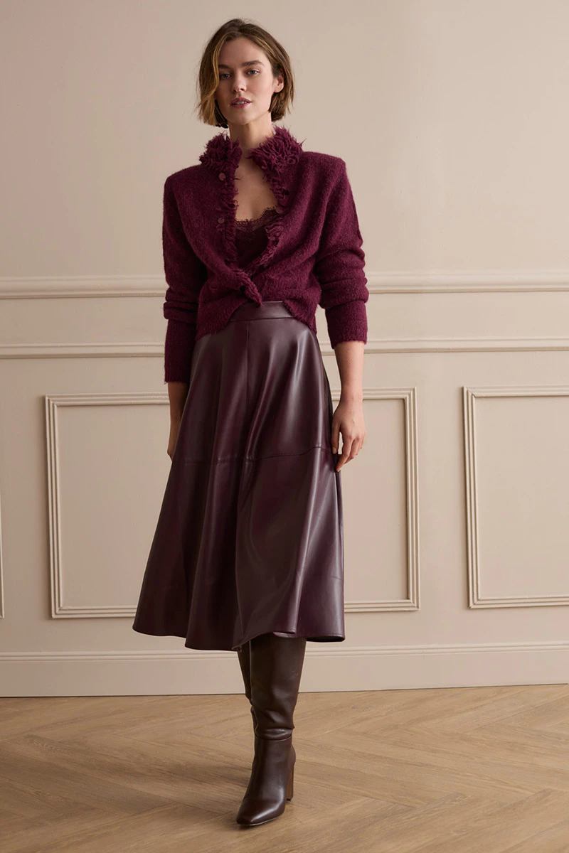 Model trägt eine burgunderfarbene Strickjacke mit Fransendetails und passendem Top zu einem dunkelroten Ledermidirock und hohen Stiefeln.