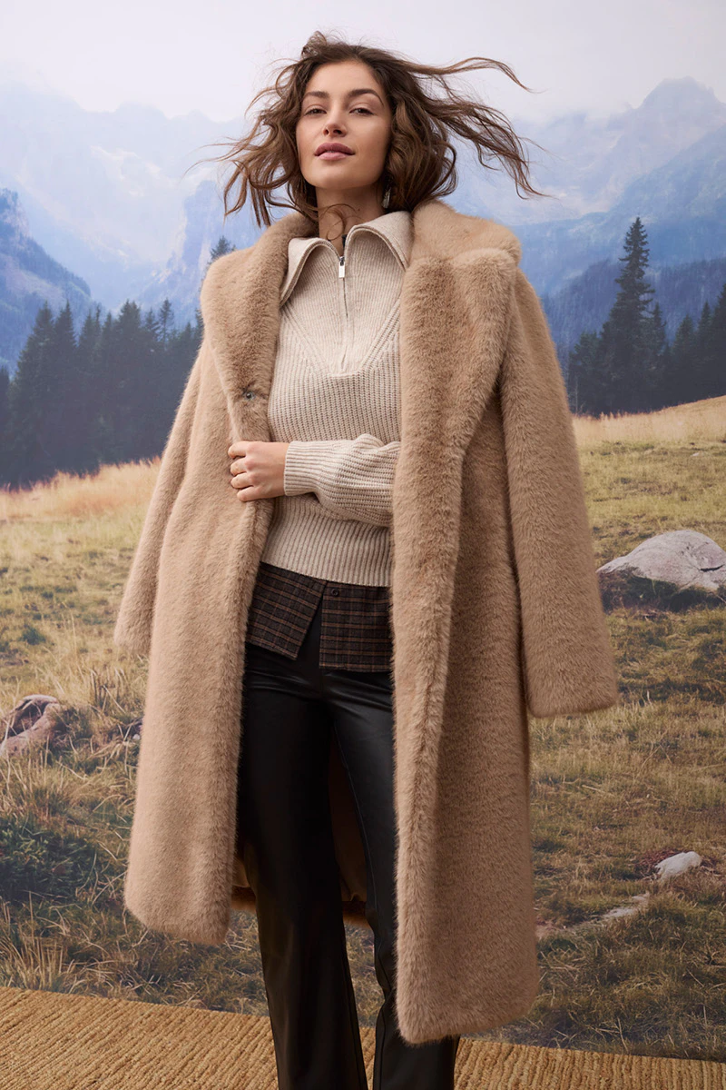 Autumn women’s fashion: a model is wearing a long beige coat over a light-coloured knitted jumper and a brown checked top, paired with black leather trousers. In the background, an alpine mountain landscape with woods and meadows can be seen.