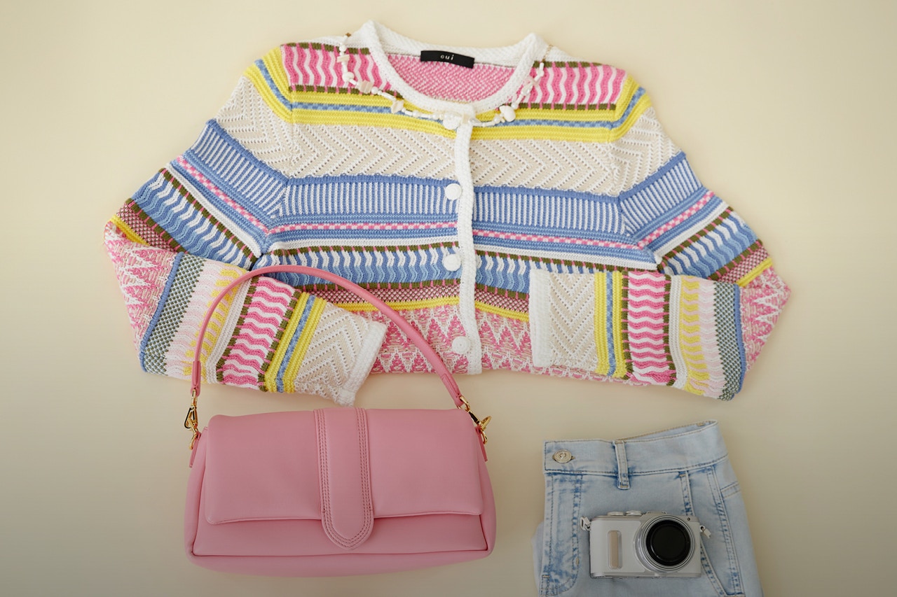 Layered fashion on a beige background. A colourful striped jacket with a pink handbag and a pair of folded jeans with a camera on top.