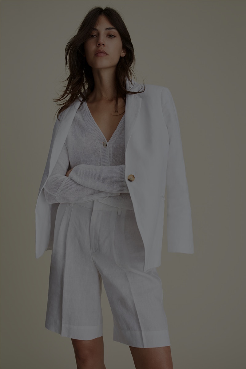 Woman wears an outfit in white. White linen blazer with a white cardigan underneath and white shorts.