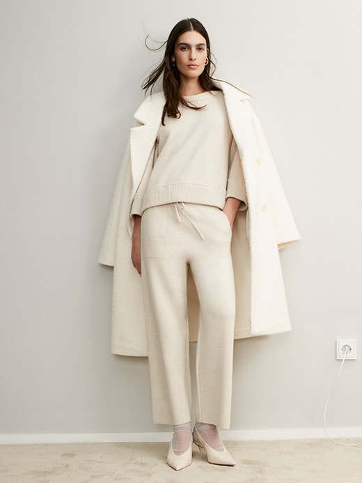 Frau an Wand gelehnt mit Händen in der weissen Hose, dazu weissen Pullover und über Schultern lässig einen weissen Mantel.