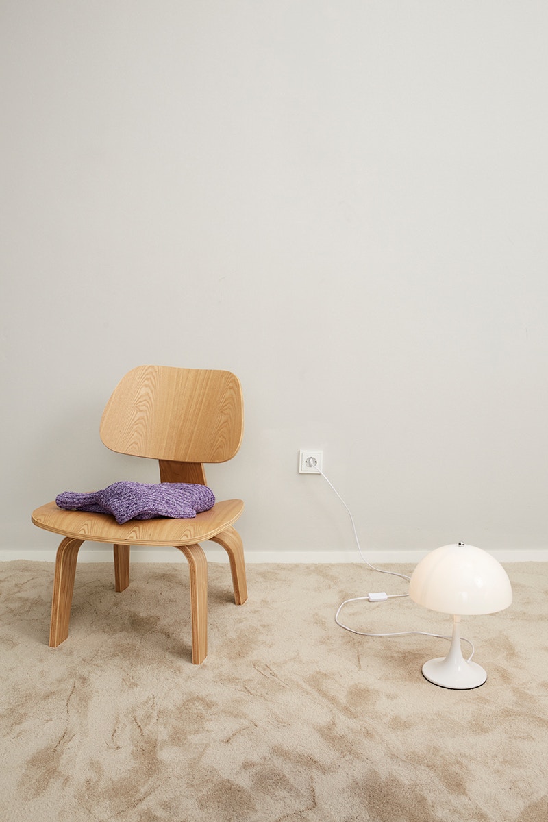 A wooden chair on the left with a purple jumper folded on top. A white lamp on the right and everything on a beige-coloured carpet.