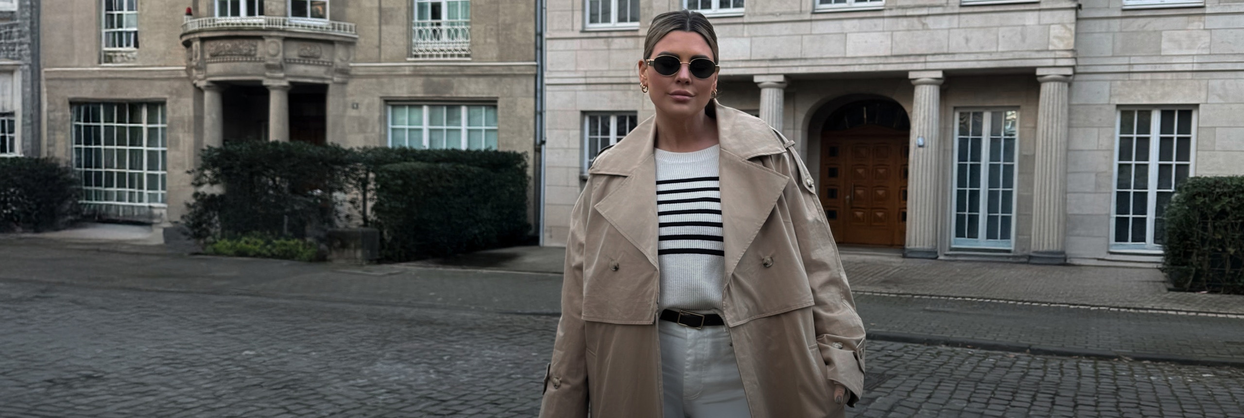 Verena Ahmann trägt den Oui Trenchcoat in lang mit einem weissen Pullover mit blauen Streifen im Oberkörper, dazu eine weisse Jeans mit weitem Bein.