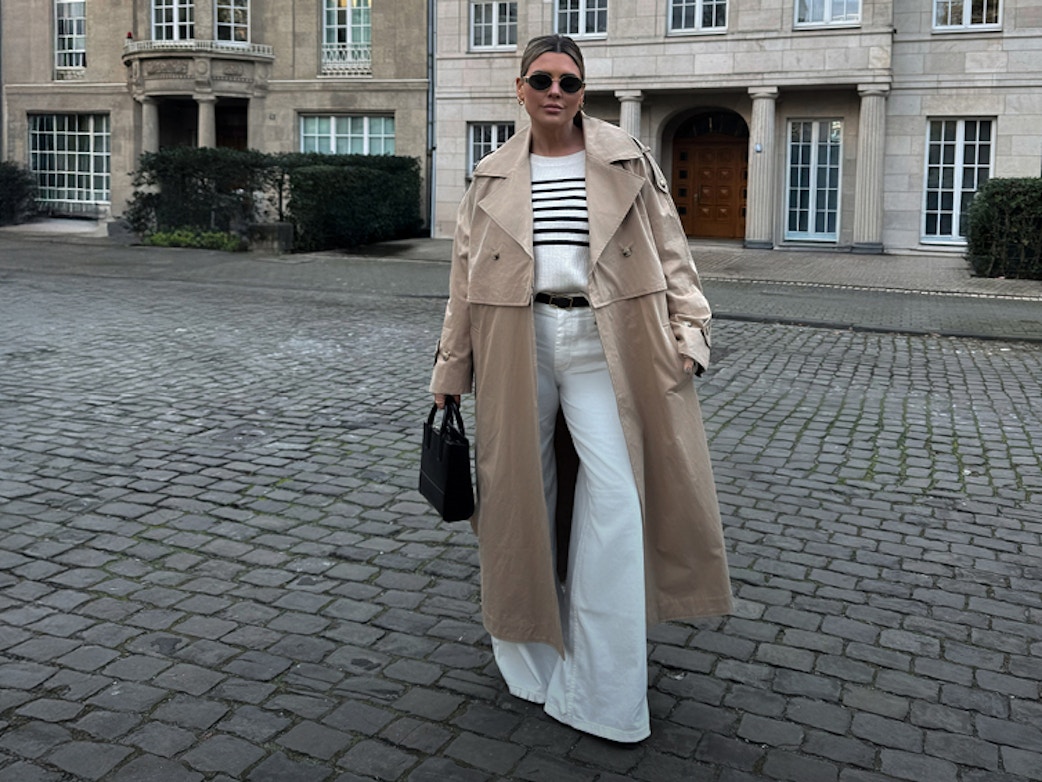 Verena Ahmann trägt den Oui Trenchcoat in lang mit einem weissen Pullover mit blauen Streifen im Oberkörper, dazu eine weisse Jeans mit weitem Bein.