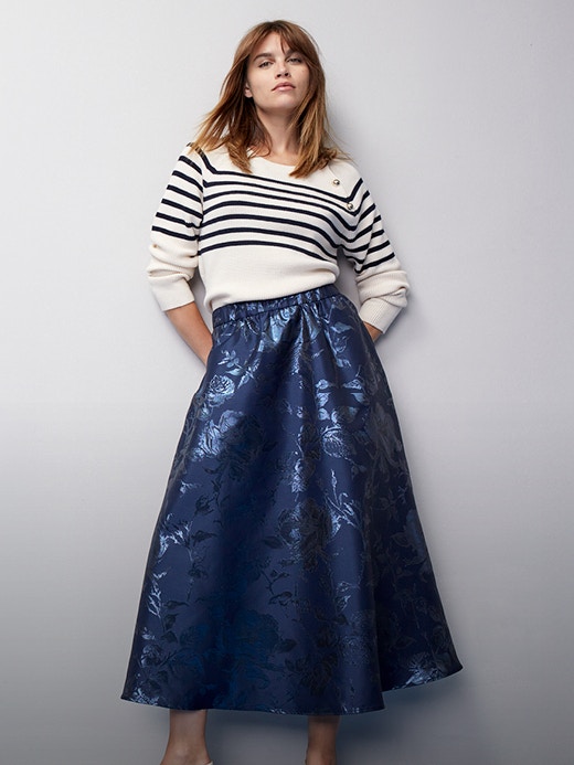 Woman wearing a white pullover with blue stripes on the upper body and a shimmering blue skirt.