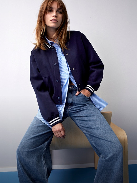 Frau mit Blouson in dunkelblau mit weissen bündchen und darunter einer hellblauen Bluse mit Jeanshose.