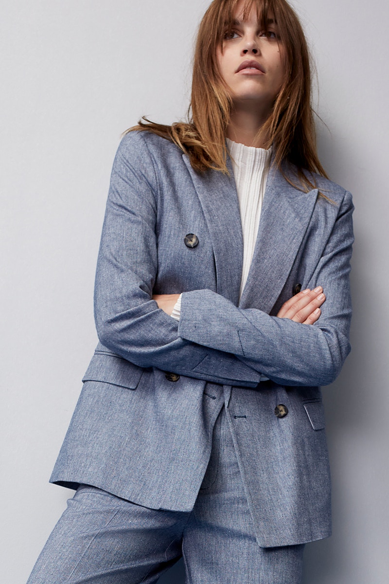 Woman wears a grey-blue suit with blazer and matching trousers and a white jumper with a slight stand-up collar underneath.