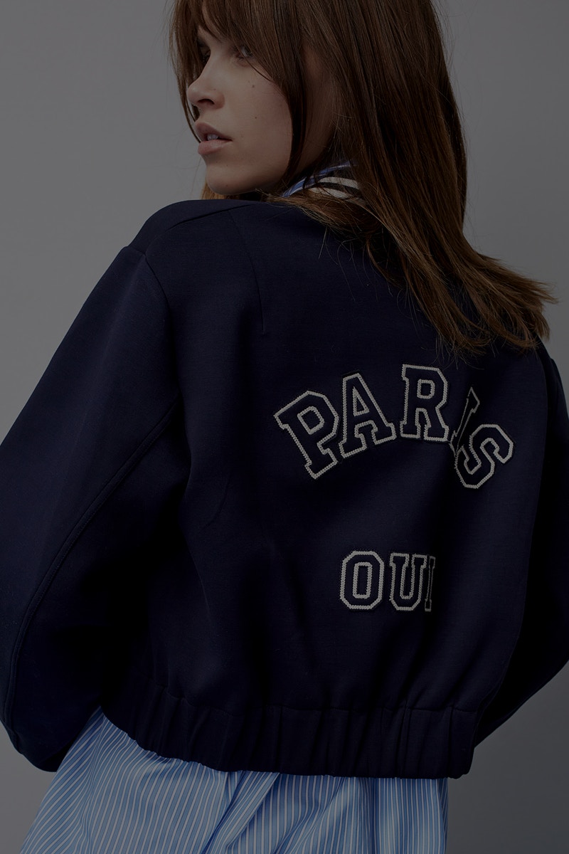 Frau mit einer dunkelblauen Blouson Jacke mit Aufdruck Paris Oui am Rücken, darunter eine hellblaues gestreiftes Hemd.