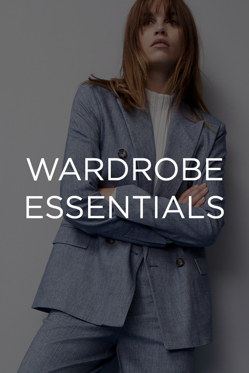 Woman wears a grey-blue suit with blazer and matching trousers and a white jumper with a slight stand-up collar underneath.