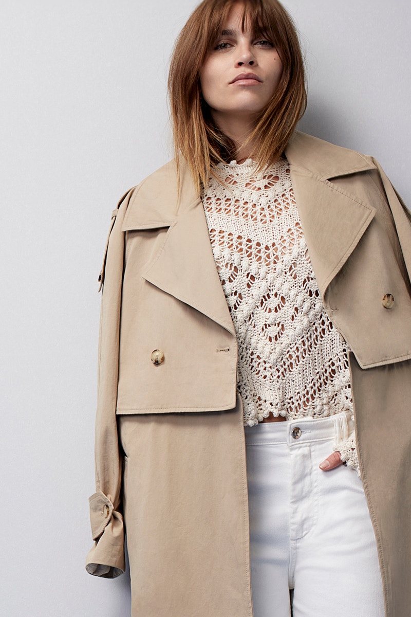 Woman with a beige trench coat and a white crocheted top with white jeans underneath.