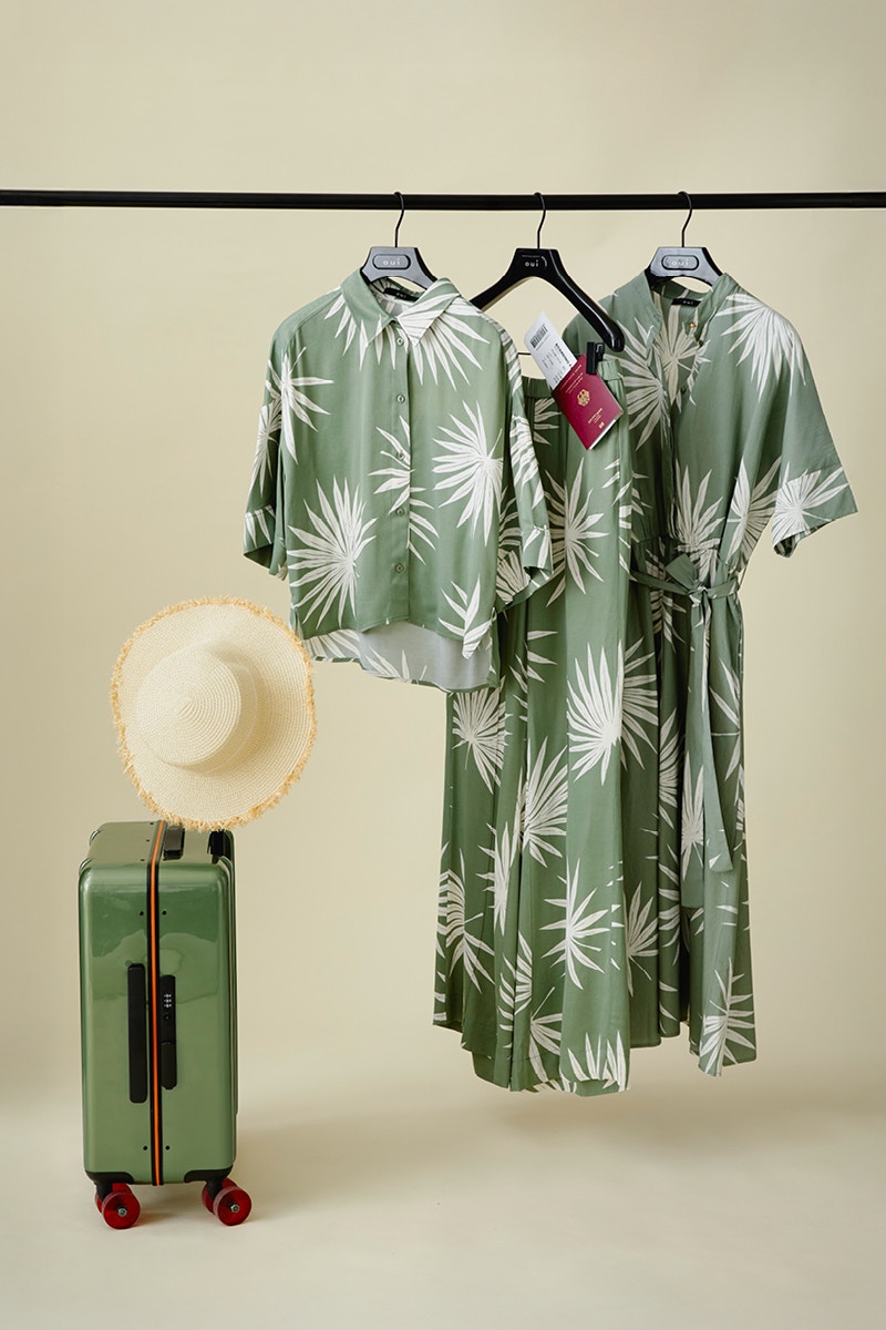 Clothes rail with blouse, skirt and dress in green with palm tree motif, in front of it a green suitcase with sun hat.