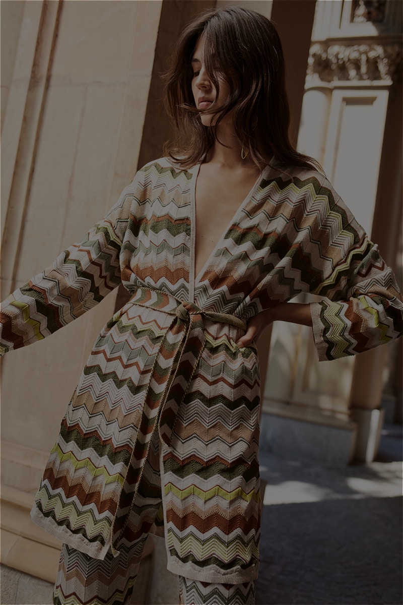 Woman with long knitted coat in zigzag pattern with various colors in green, brown, white and beige.