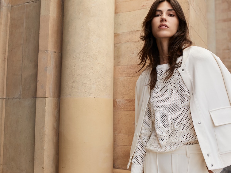 Woman wears a white jacket with a white knitted jumper in a crocheted look with palm motifs and matching white trousers.