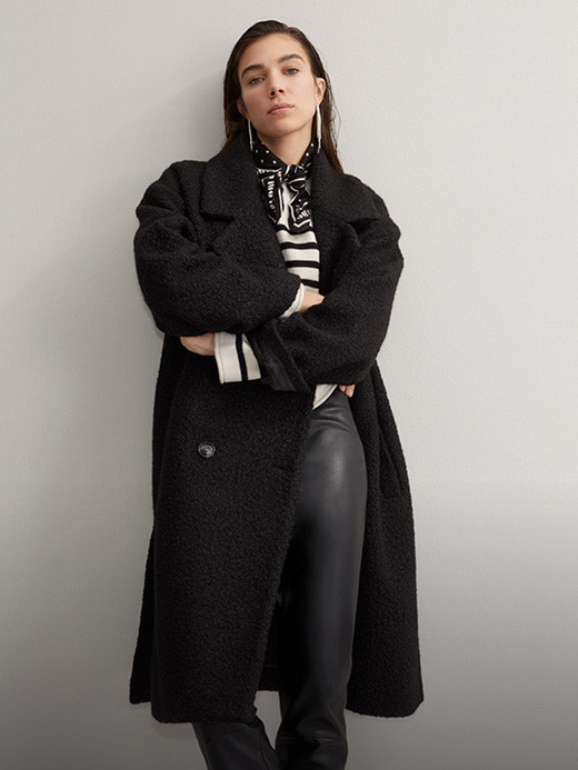 A woman leans casually against a grey wall. She is wearing an outfit with a black knee-length coat, black trousers and a black and white striped jumper. Click on the picture to buy the look directly.