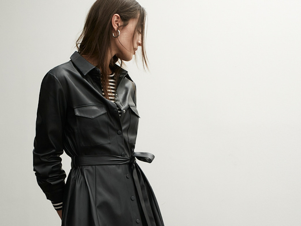 Young woman stands in a studio setting looking to the right with her arms behind her back. She is wearing a black leather dress with a tie ribbon and a black and white striped top peeking out from the neckline.