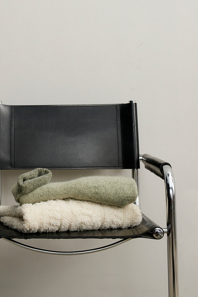 A metal chair with a black back and seat. Two items of clothing are draped over it. The top one in green and a knitted jumper in white underneath.