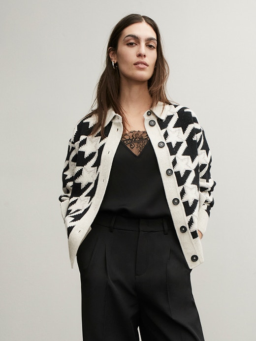 Young woman with her hands in her pockets wearing a white and black patterned cardigan. Underneath is a black top with a lace insert at the neckline and black trousers.