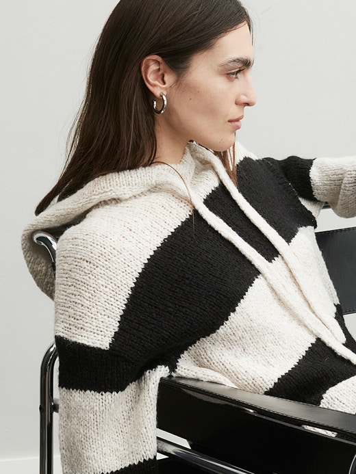 Young woman sitting casually in a chair wearing a knitted jumper with hood with block stripes in white and black.
