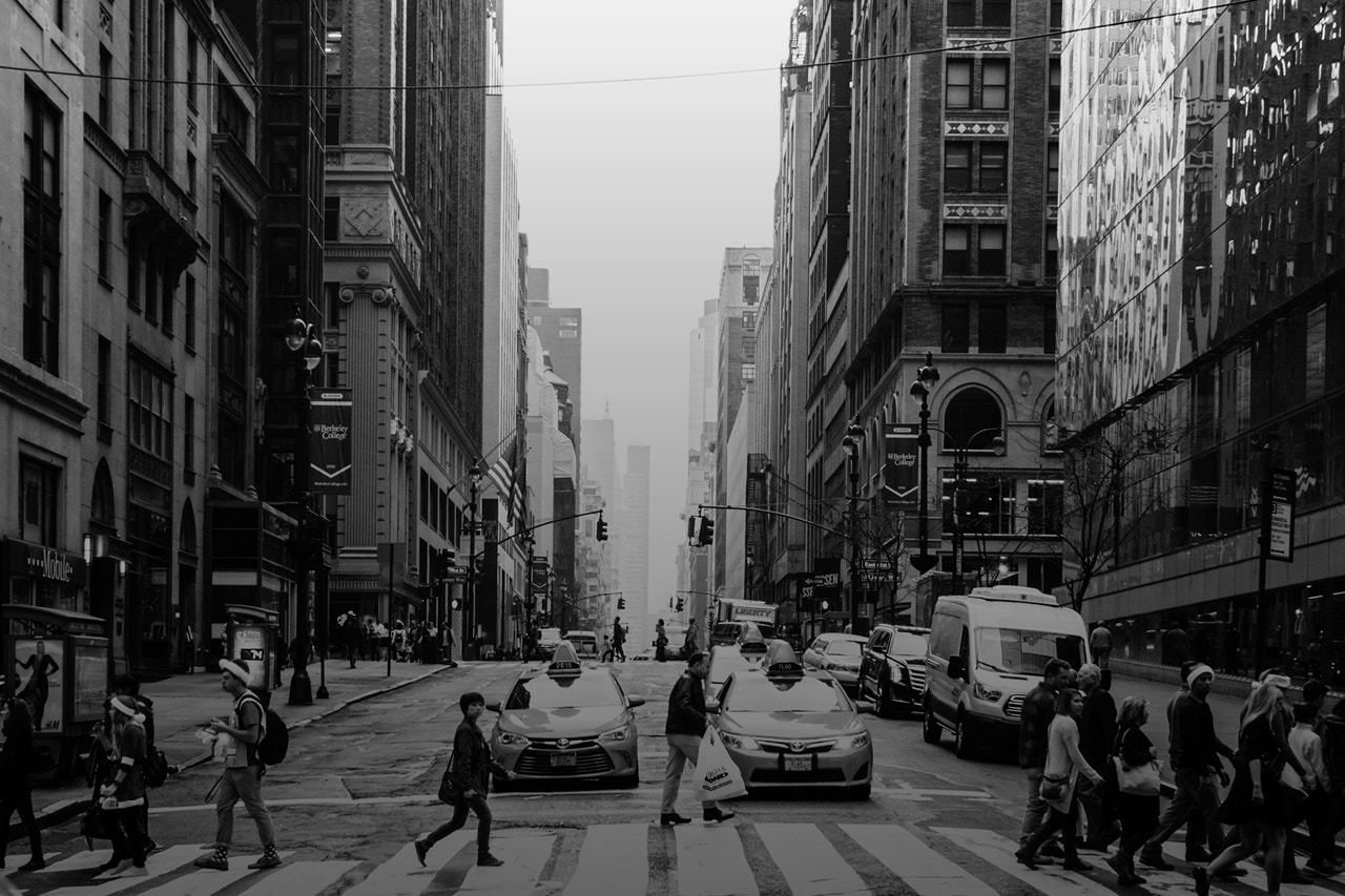 A large street with people walking across the zebra crossing. There are tall buildings to the left and right. The picture is in black and white and taken in New York.