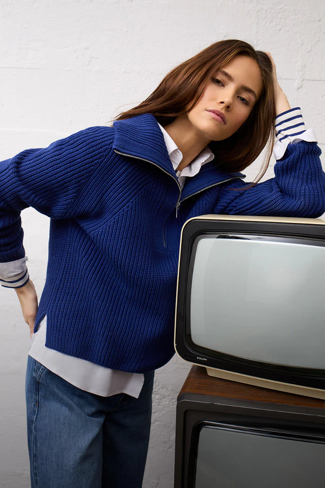 Frau im blauen Strickpullover mit Reißverschluss und weißem Hemd mit gestreiften Manschetten, lässig an einem Retro-Fernseher gelehnt.
