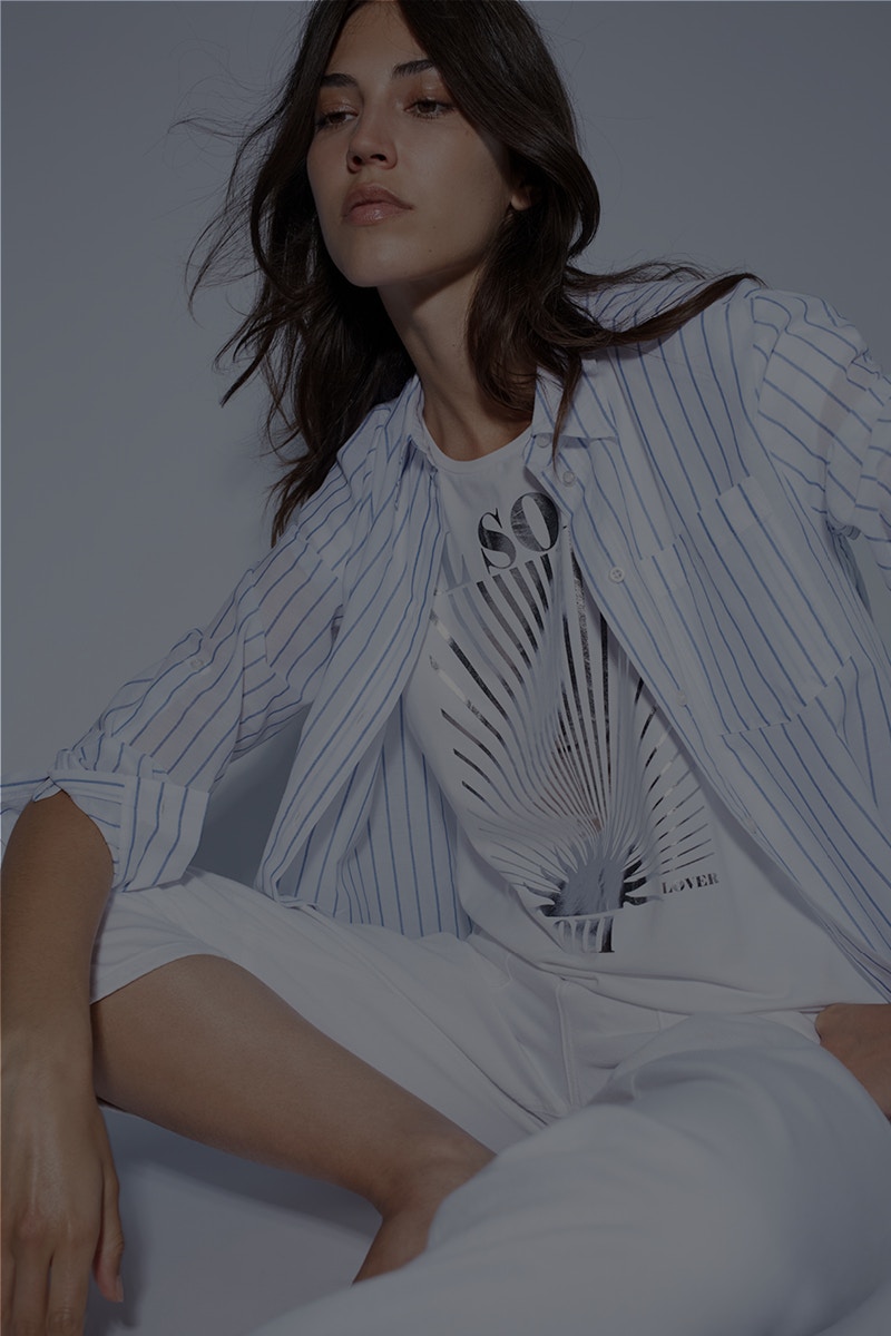 Woman wearing a striped white and blue blouse underneath a shirt with a front print and short white trousers.