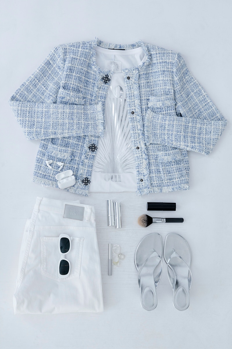 A picture of folded clothes. A jacket in light blue chequered with a top in white underneath, on the left a pair of folded white jeans with sunglasses in the pocket, on the right silver sandals and some make-up accessories.