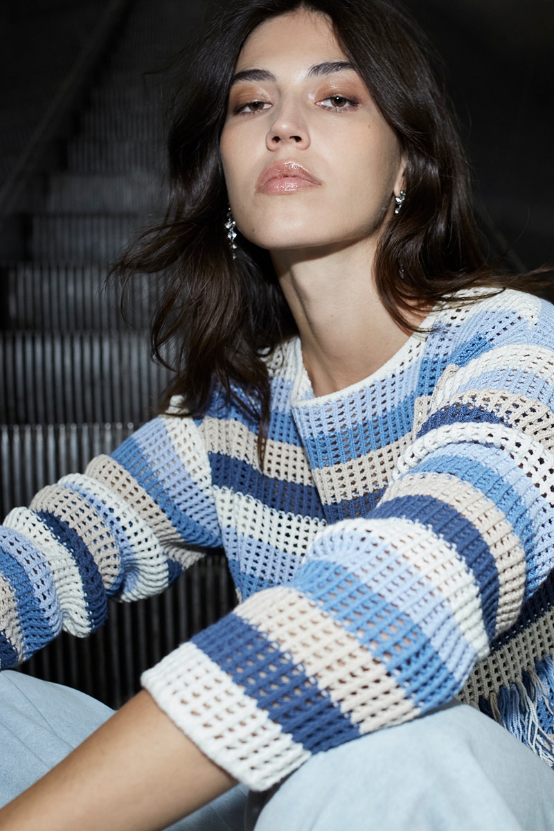 Woman wears a crocheted jumper in different block stripes of blue, white and beige and a pair of light blue jeans.
