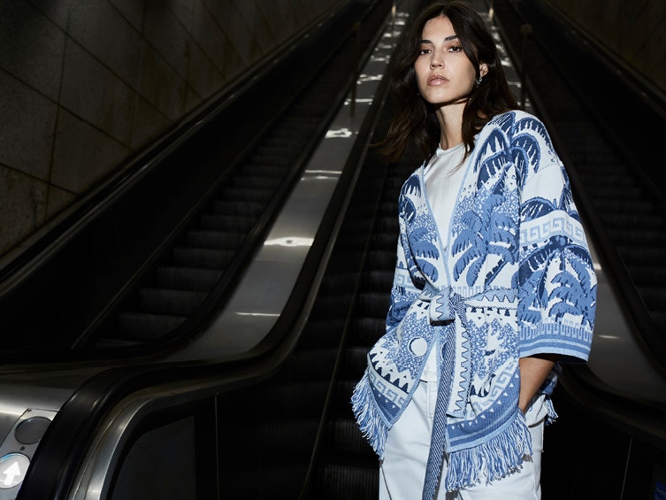 Woman wears a cardigan with palm jacquard in white and blue and white trousers.